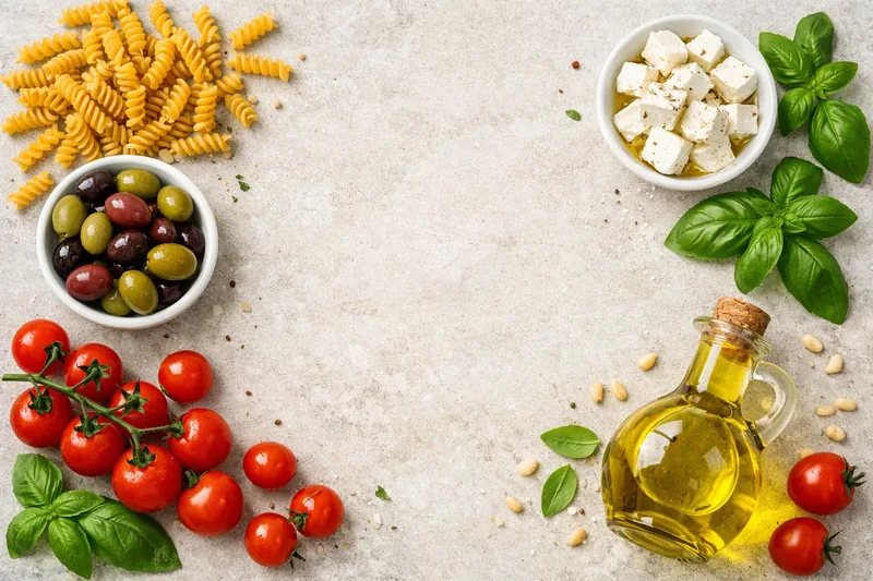 Zutaten für Nudelsalat auf hellem Stein: Spirelli, eine Schale Oliven, Feta, Basilikum, Kirschtomaten und eine Flasche Olivenöl.